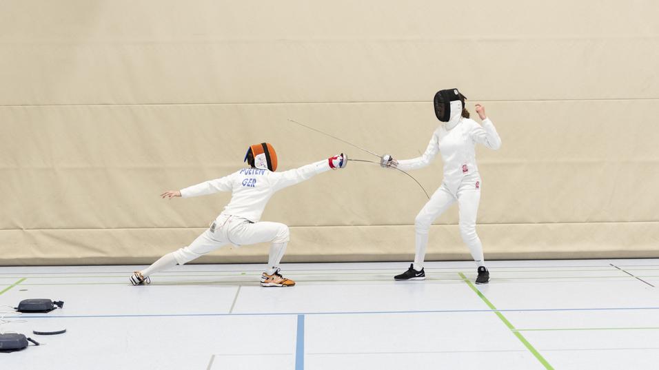 Zwei Fechter in Sportbekleidung k&auml;mpfen in einer Turnhalle; einer greift an, der andere weicht aus.