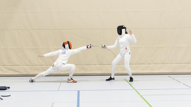 Zwei Fechter in Sportbekleidung k&auml;mpfen in einer Turnhalle; einer greift an, der andere weicht aus.
