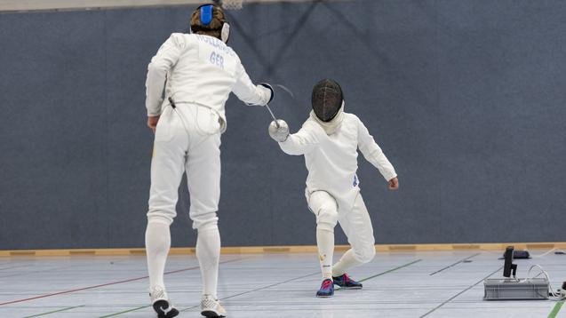 Zwei Fechter in wei&szlig;er Ausr&uuml;stung k&auml;mpfen in einer Sporthalle auf einem gepflasterten Boden.