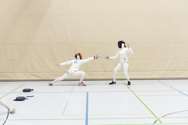 Zwei Fechter in Sportbekleidung k&auml;mpfen in einer Turnhalle; einer greift an, der andere weicht aus.