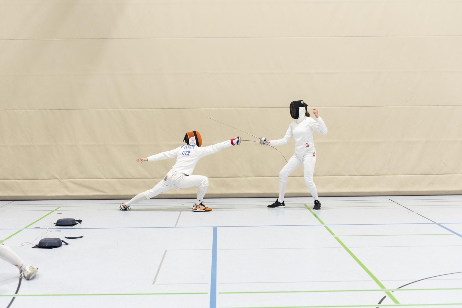 Zwei Fechter in Sportbekleidung k&auml;mpfen in einer Turnhalle; einer greift an, der andere weicht aus.