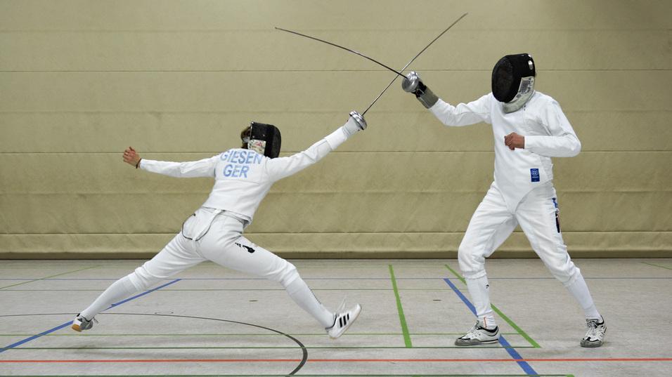 Zwei Fechter in Sportkleidung k&auml;mpfen mit Floretten in einer Halle, w&auml;hrend sie sich aktiv gegen&uuml;berstehen.