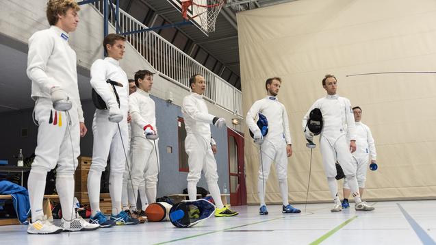 Sechs Fechter in Wettkampfanz&uuml;gen stehen in einer Sporthalle, bereit f&uuml;r das Training.