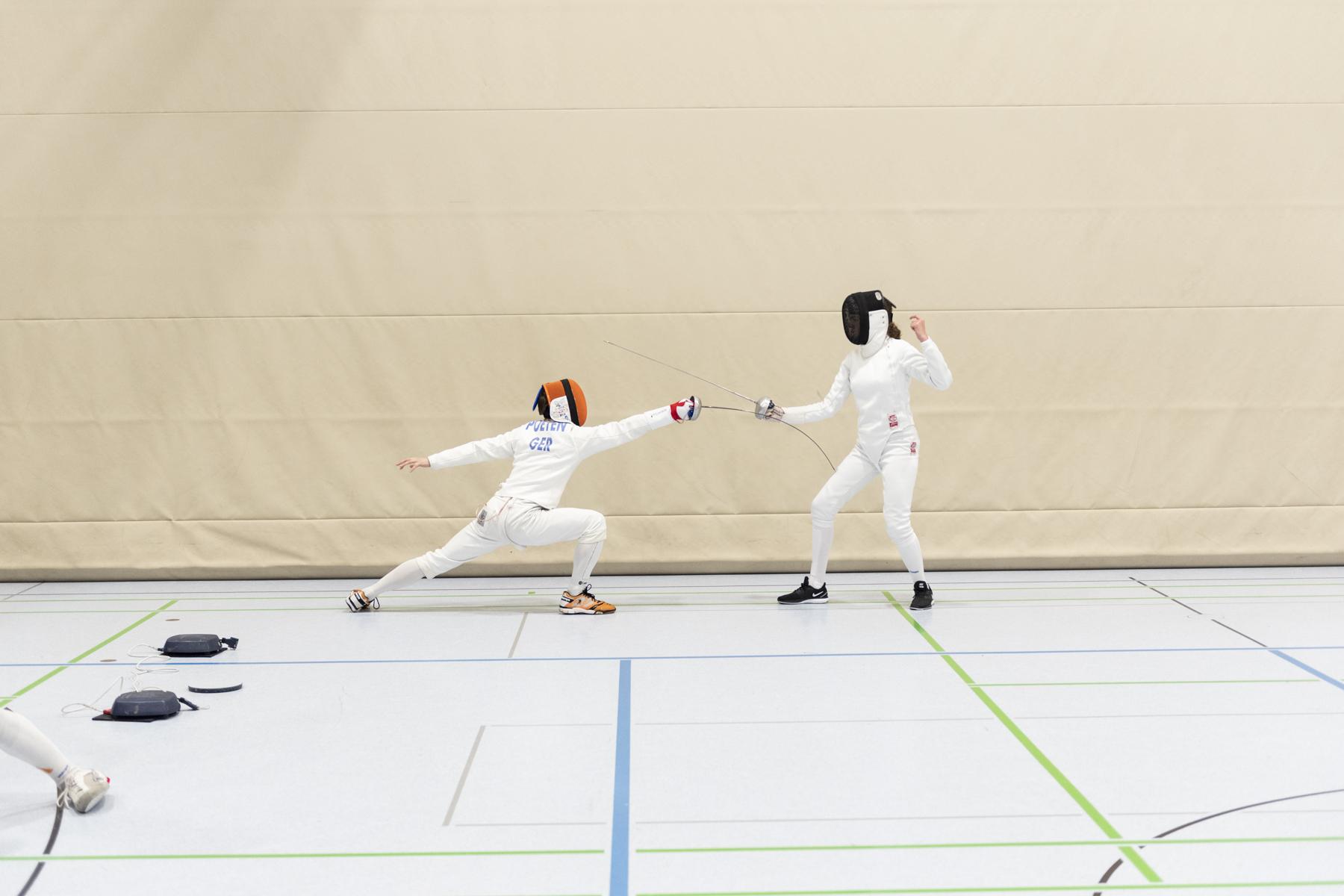Zwei Fechter in Sportbekleidung k&auml;mpfen in einer Turnhalle; einer greift an, der andere weicht aus.