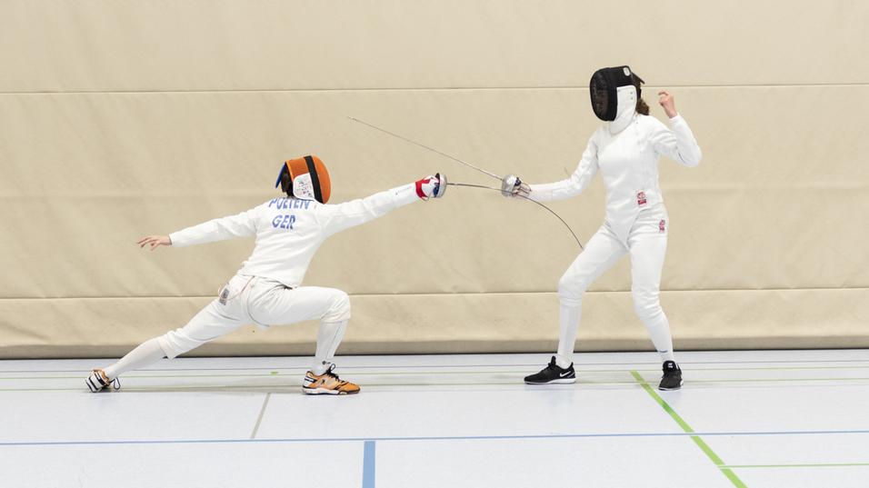 Zwei Fechter in Sportbekleidung k&auml;mpfen in einer Turnhalle; einer greift an, der andere weicht aus.