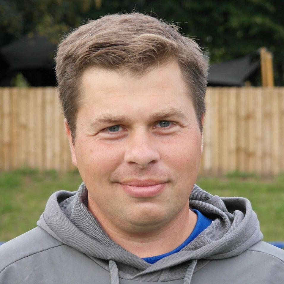 Porträt eines Mannes mit kurzem, braunem Haar, in grauem Sweatshirt, vor einem unscharfen, natürlichen Hintergrund.