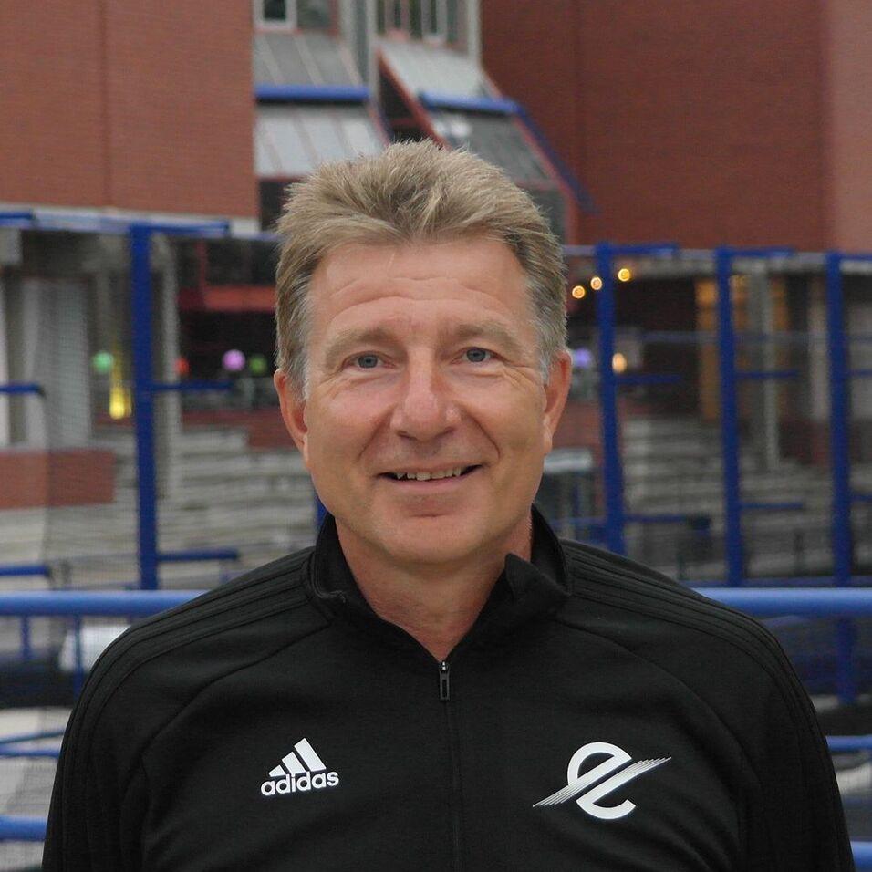 Lächelnder Mann mit kurzen, grauen Haaren trägt ein schwarzes Sportoberteil mit Logo, Hintergrund ist ein Sportgelände.