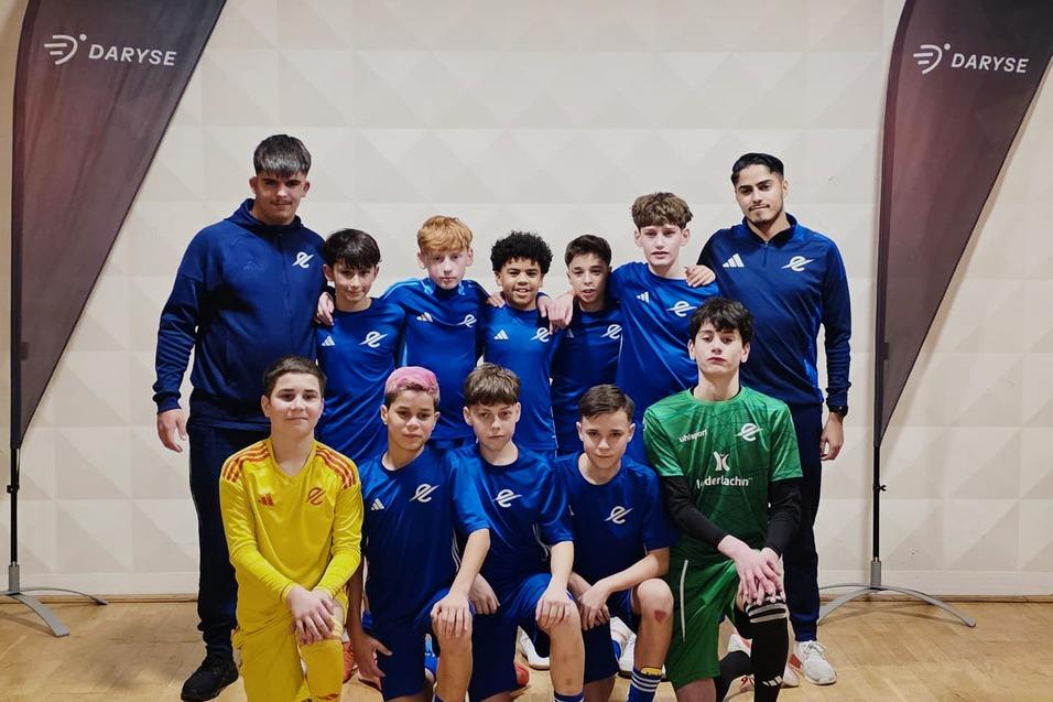 Jugendfu&szlig;ballmannschaft mit Trainer in blauen Trikots, posierend vor DARYSE-Flaggen in einer Sporthalle.