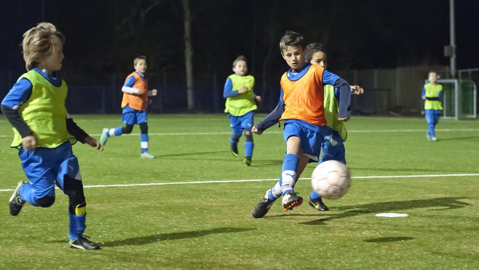 Kinder im Fu&szlig;balltraining spielen abends auf einem Kunstrasenplatz, w&auml;hrend einer den Ball passt.