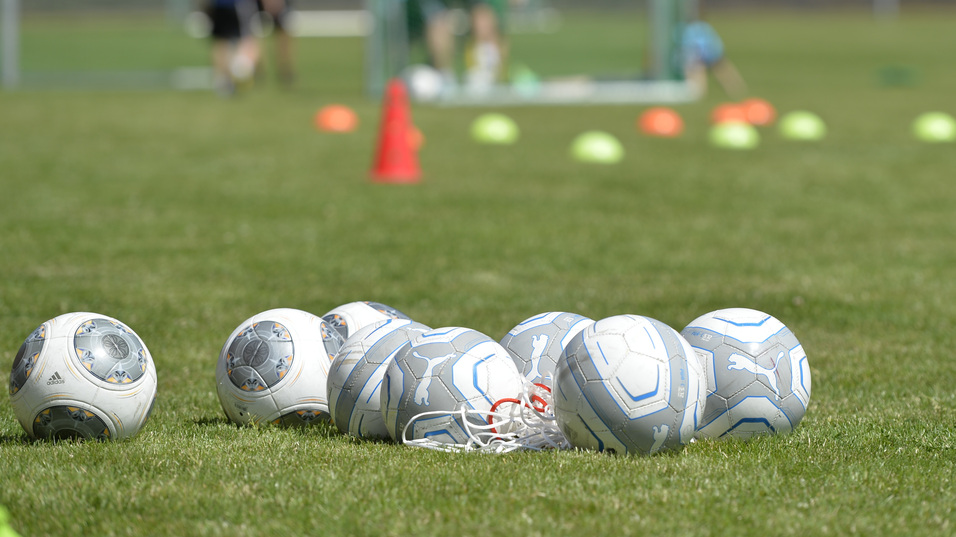 Mehrere Fu&szlig;ballb&auml;lle liegen auf einem Rasenplatz, im Hintergrund trainieren Spieler vor zwei Toren.