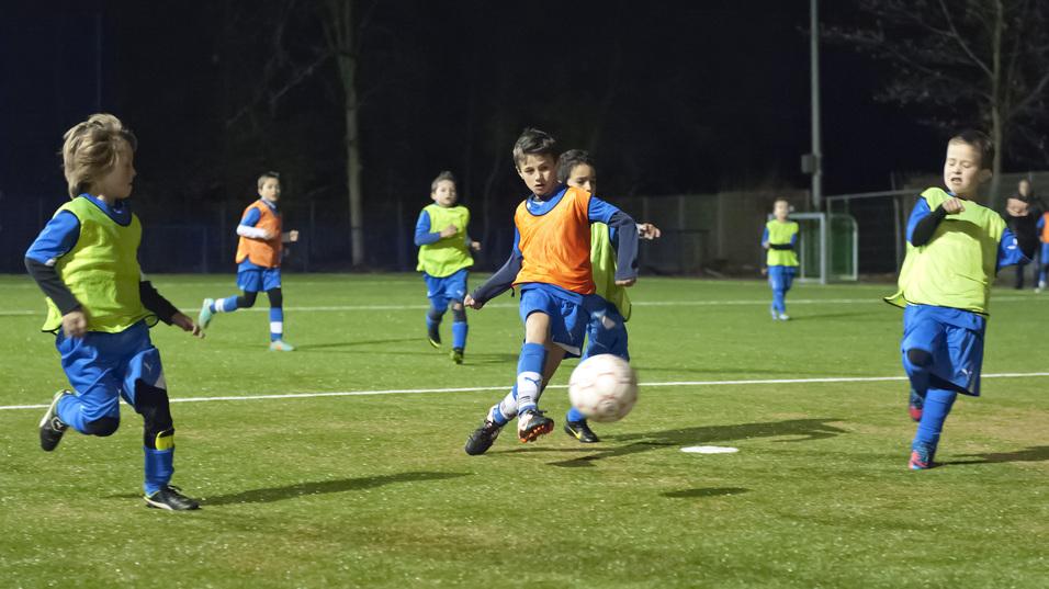 Kinder im Fu&szlig;balltraining spielen abends auf einem Kunstrasenplatz, w&auml;hrend einer den Ball passt.