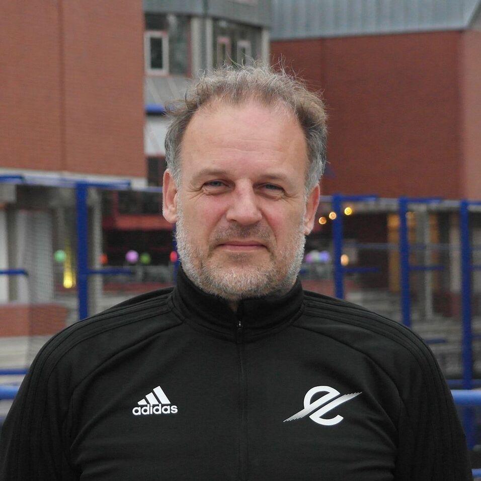 Ein Mann mit grauen Haaren und Bart trägt einen schwarzen Adidas-Jacket und steht vor einer urbanen Kulisse.