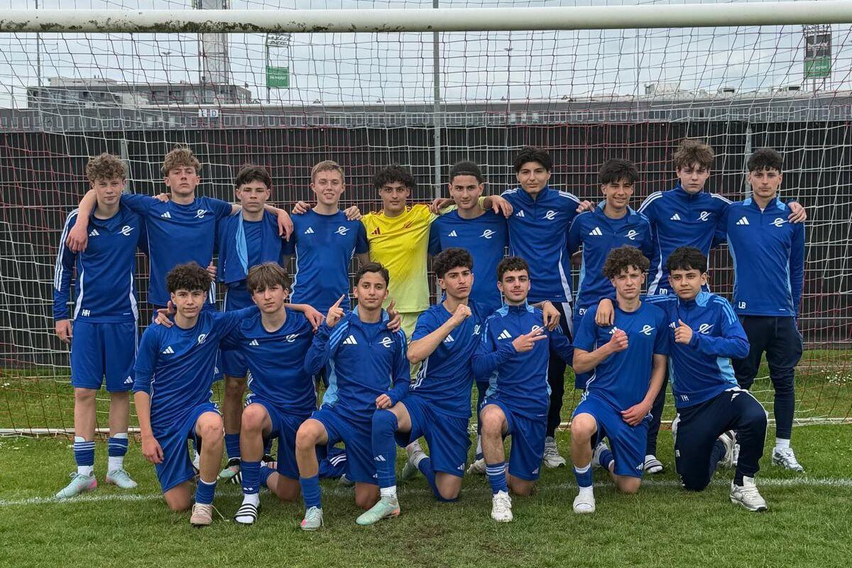 Gruppe von 15 jungen Fußballspielern in blauen Trikots steht vor einem Fußballtor auf einem Rasenfeld.