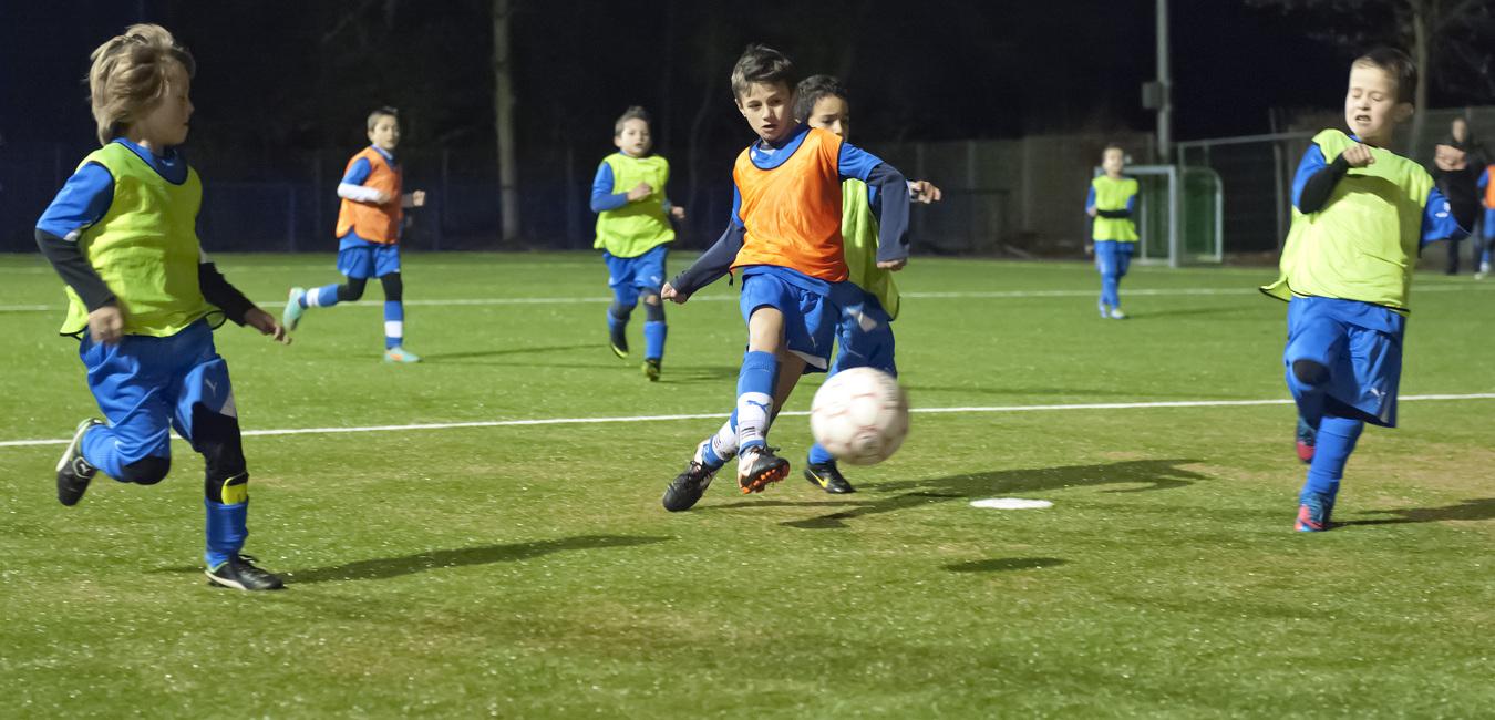 Kinder im Fußballtraining spielen abends auf einem Kunstrasenplatz, während einer den Ball passt.
