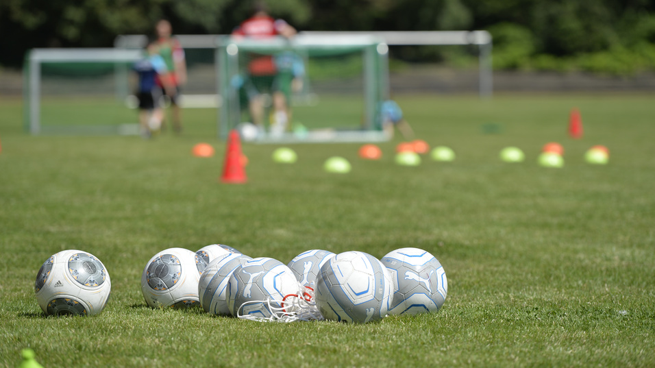 Mehrere Fu&szlig;ballb&auml;lle liegen auf einem Rasenplatz, im Hintergrund trainieren Spieler vor zwei Toren.