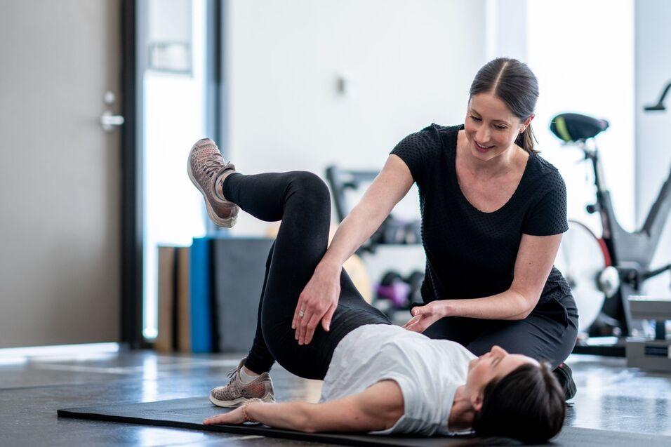 Zwei Frauen in Sportkleidung üben in einem Fitnessstudio, eine Trainerin hilft der anderen bei einer Übung auf der Matte.