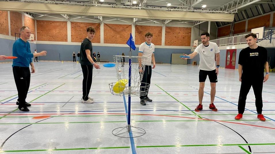 Fünf junge Männer stehen in einer Sporthalle, bereit, mit Frisbees in einen Korb zu werfen.