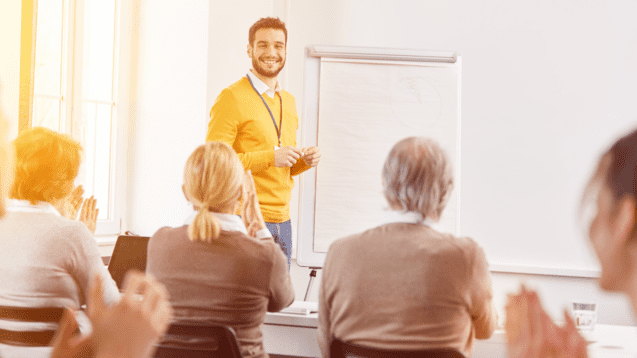 Ein lächelnder Mann in einem gelben Pullover steht vor einem Whiteboard, während Teilnehmer ihm applaudieren.