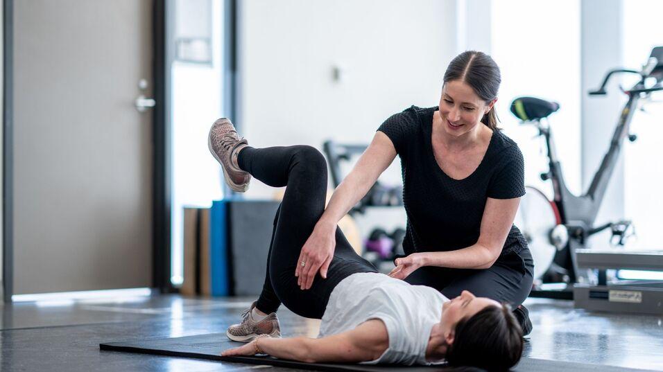 Zwei Frauen in Sportkleidung &uuml;ben in einem Fitnessstudio, eine Trainerin hilft der anderen bei einer &Uuml;bung auf der Matte.