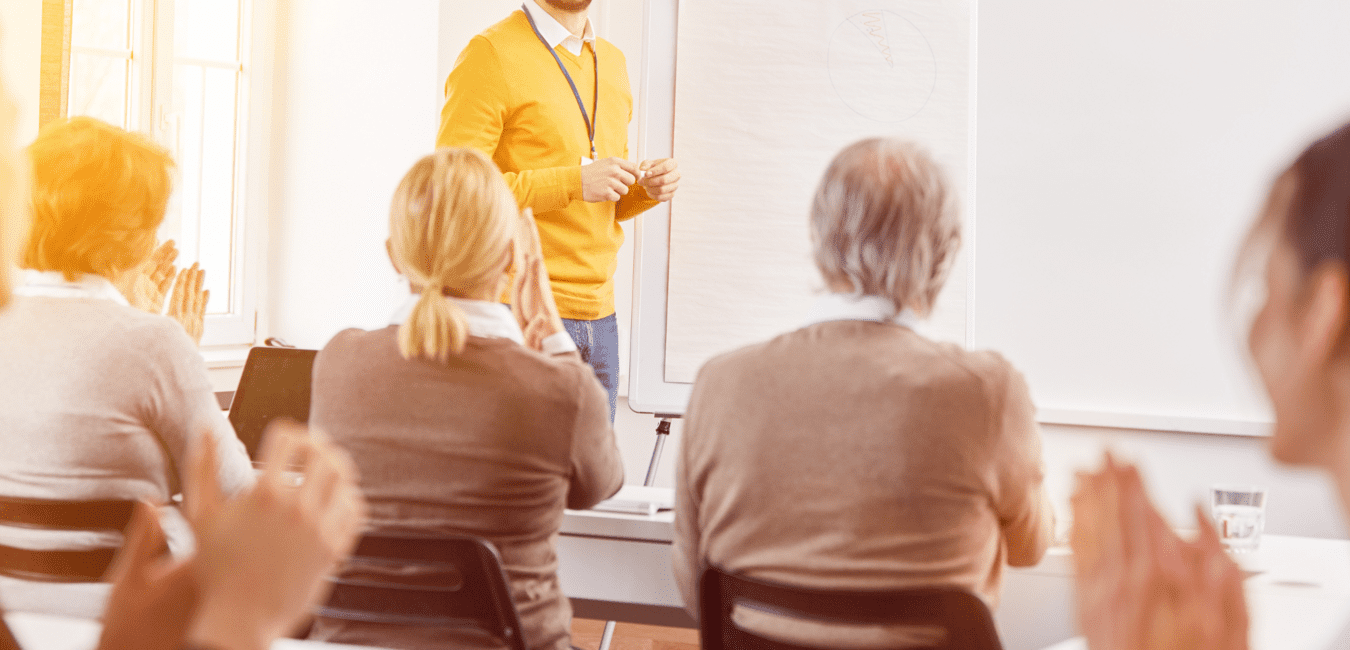 Ein lächelnder Mann in einem gelben Pullover steht vor einem Whiteboard, während Teilnehmer ihm applaudieren.