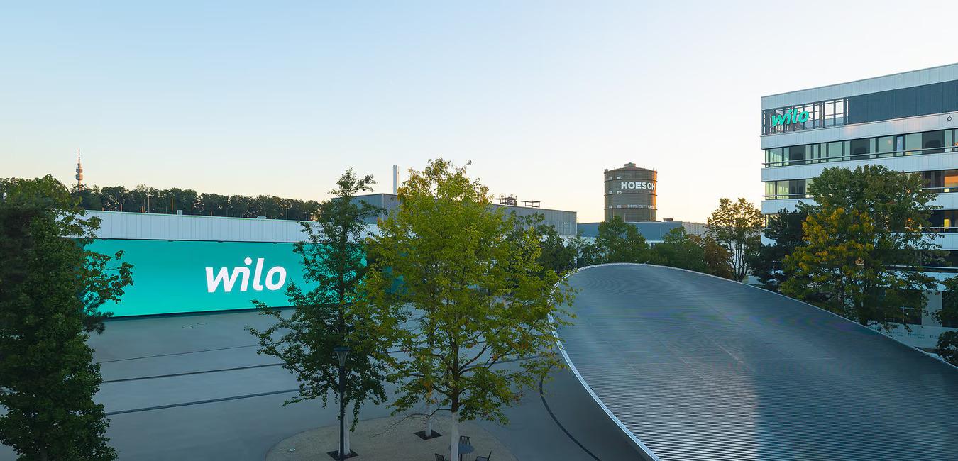 Stadtansicht mit dem Schriftzug &bdquo;wilo&ldquo; auf einem Geb&auml;ude und B&auml;umen im Vordergrund bei Sonnenaufgang.