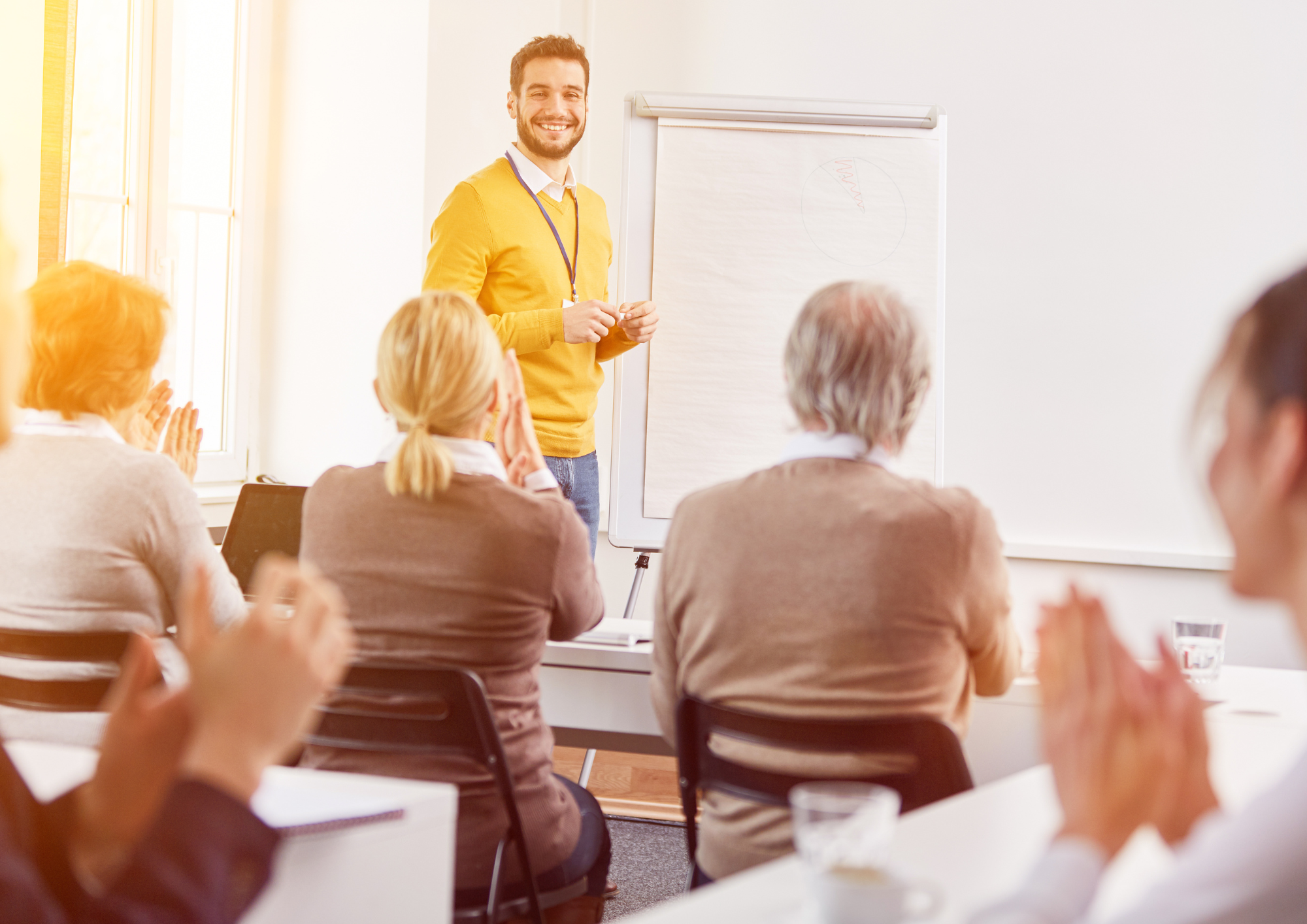 Ein lächelnder Mann in einem gelben Pullover steht vor einem Whiteboard, während Teilnehmer ihm applaudieren.