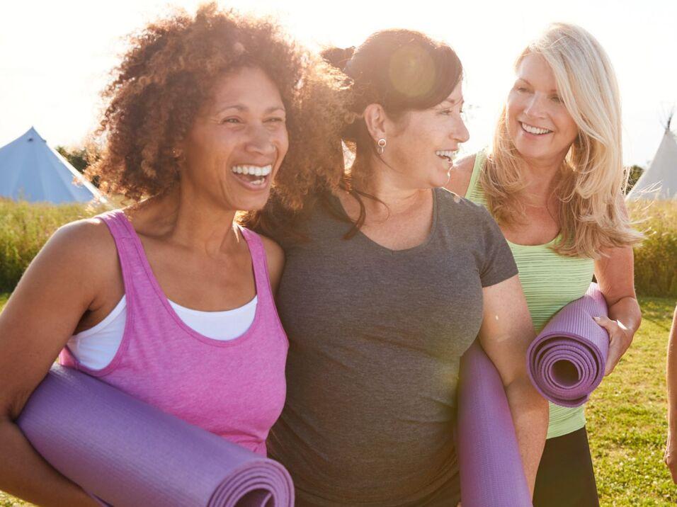 Vier Frauen mit Yogamatten in der Hand lachen und unterhalten sich auf einer Wiese mit Zelten im Hintergrund.