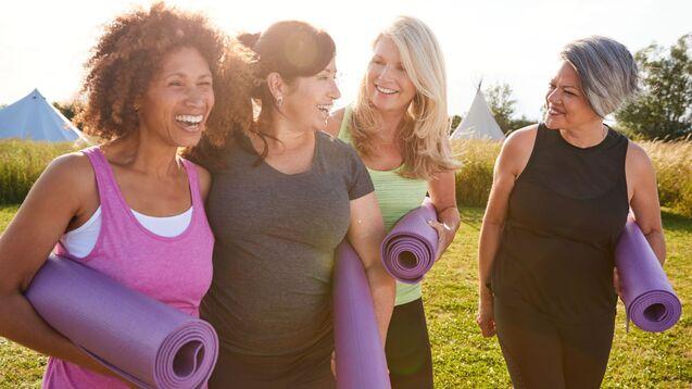Vier Frauen mit Yogamatten in der Hand lachen und unterhalten sich auf einer Wiese mit Zelten im Hintergrund.