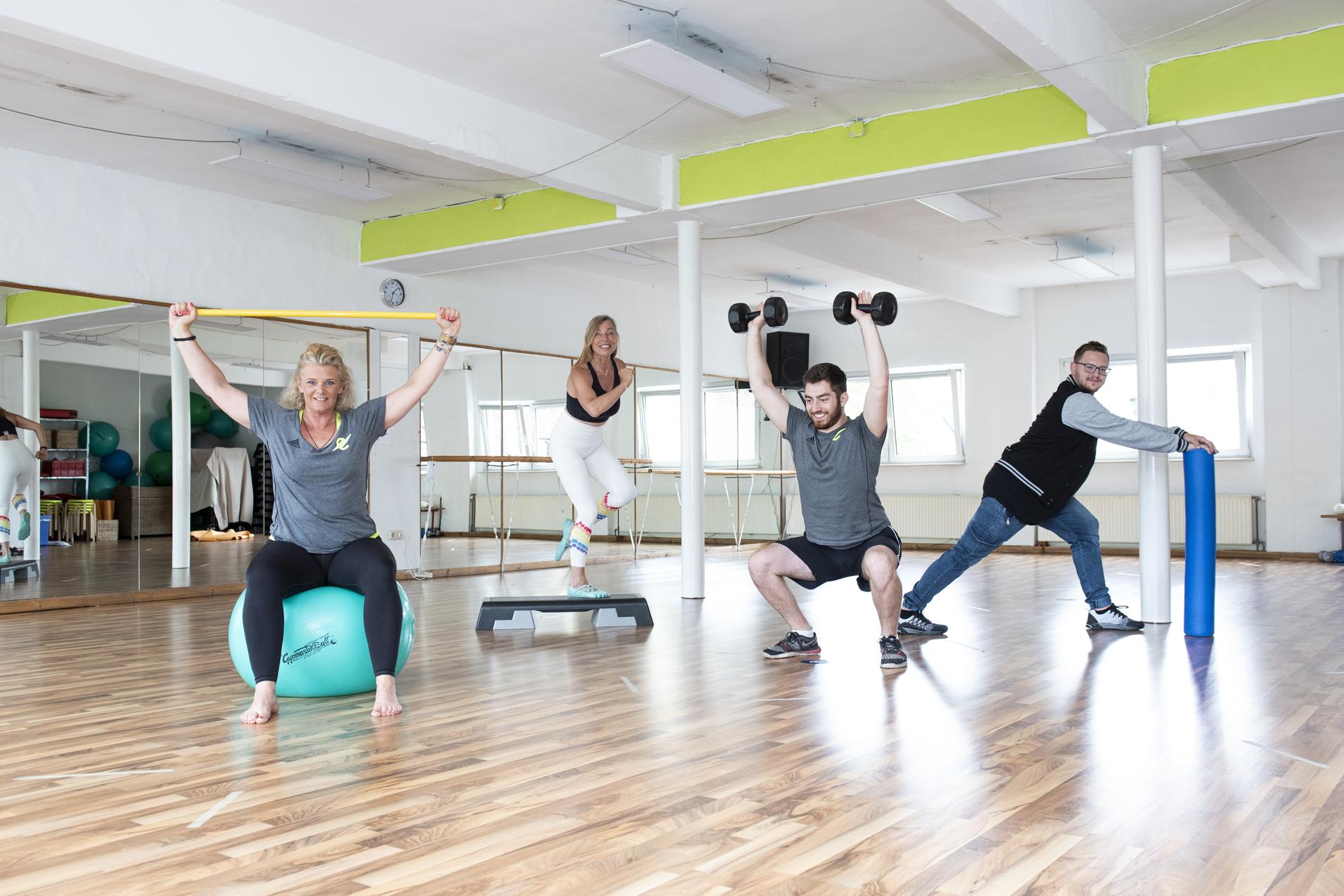 Vier Personen trainieren in einem Fitnessraum: Eine sitzt auf einem Gymnastikball, zwei heben Gewichte, eine dehnt sich.