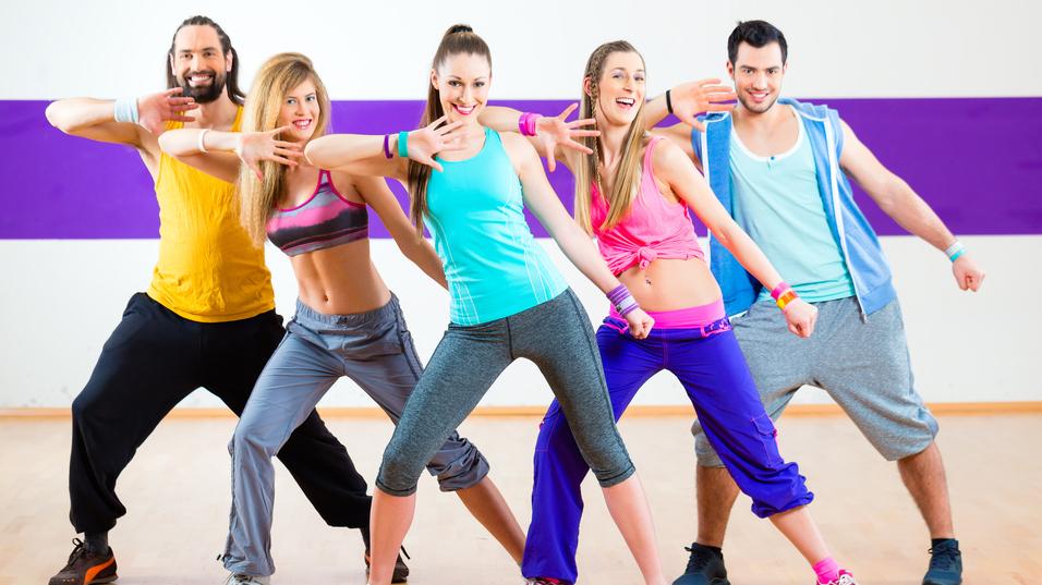 F&uuml;nf tanzende Personen in Sportkleidung zeigen synchron eine Pose in einem Fitnessraum mit farbiger Wand.