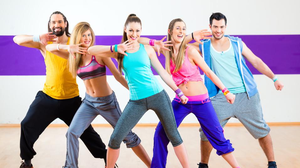 Fünf tanzende Personen in Sportkleidung zeigen synchron eine Pose in einem Fitnessraum mit farbiger Wand.