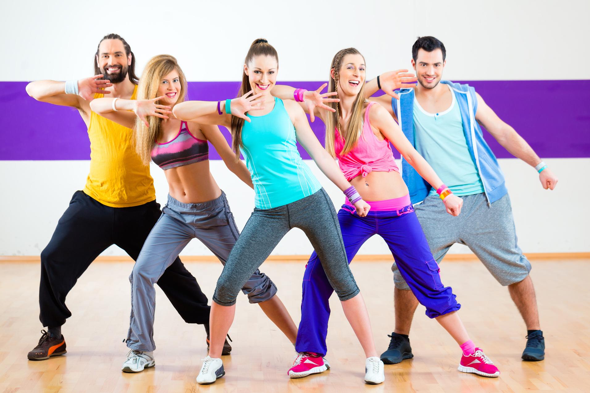 F&uuml;nf tanzende Personen in Sportkleidung zeigen synchron eine Pose in einem Fitnessraum mit farbiger Wand.