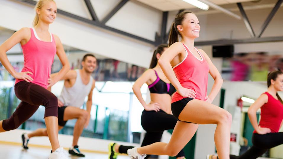Gruppe von f&uuml;nf Personen in Sportkleidung beim Training in einem Fitnessstudio, Schwerpunkt auf Ausfallschritten.