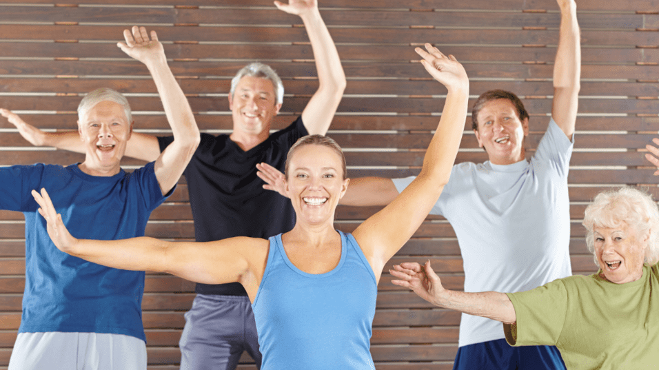 Gruppe älterer Erwachsenen macht gemeinsam Übungen in einem Fitnesskurs mit fröhlichen Gesichtern und gestreckten Armen.