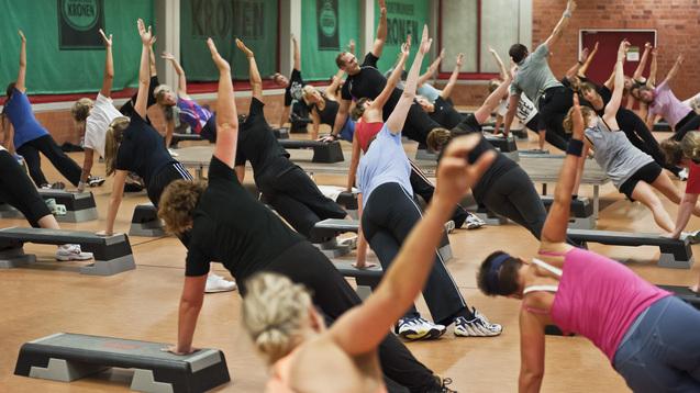 Gruppe von Fitnessübenden in einer Sporthalle, die gemeinsam Dehn- und Kräftigungsübungen durchführen.