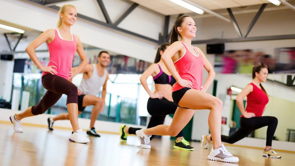 Gruppe von fünf Personen in Sportkleidung beim Training in einem Fitnessstudio, Schwerpunkt auf Ausfallschritten.