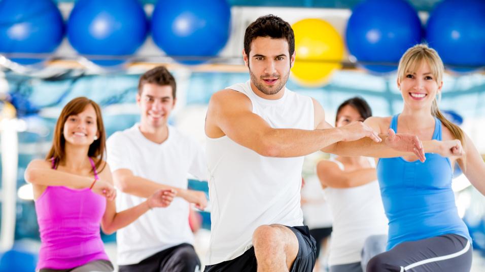 Eine Gruppe von f&uuml;nf Personen macht Fitness&uuml;bungen in einem Studio mit bunten Gymnastikb&auml;llen im Hintergrund.