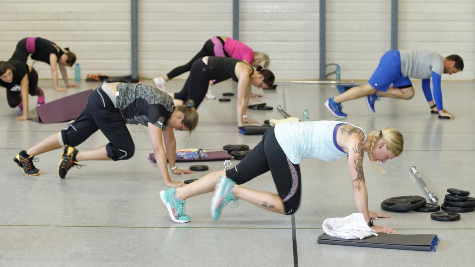 Gruppe von Personen in Sportkleidung beim Training mit Klimmzügen und Gewichten in einer Turnhalle.