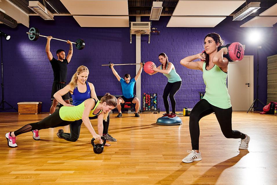 Gruppenfitness: Sechs Personen trainieren gemeinsam im Gym mit Gewichten und Fitnessgeräten auf einem Holzboden.