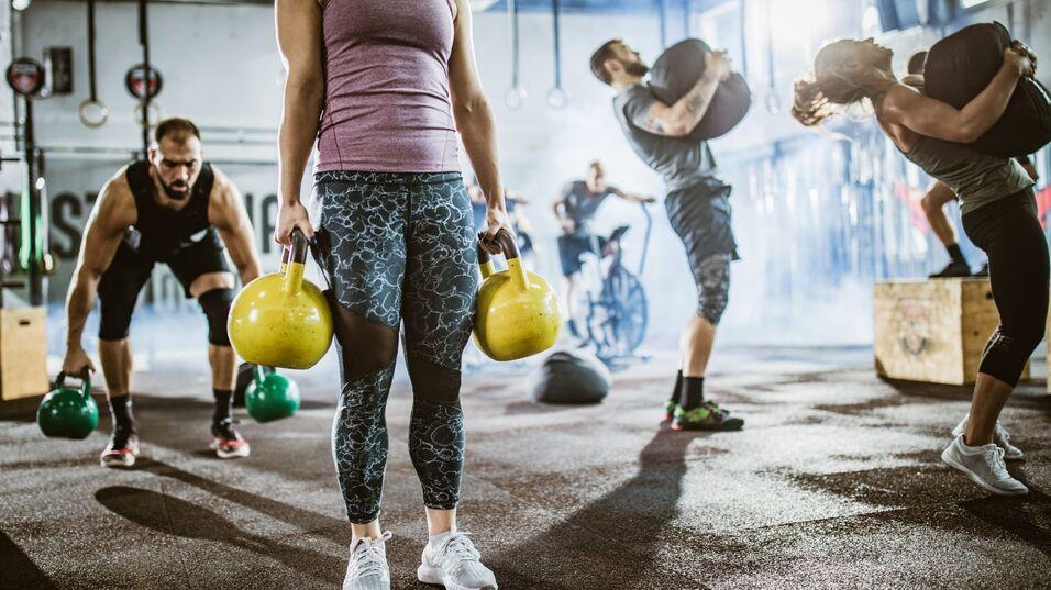 Person mit Kettlebells im Vordergrund, während andere im Hintergrund verschiedene Übungen im Fitnessstudio ausführen.