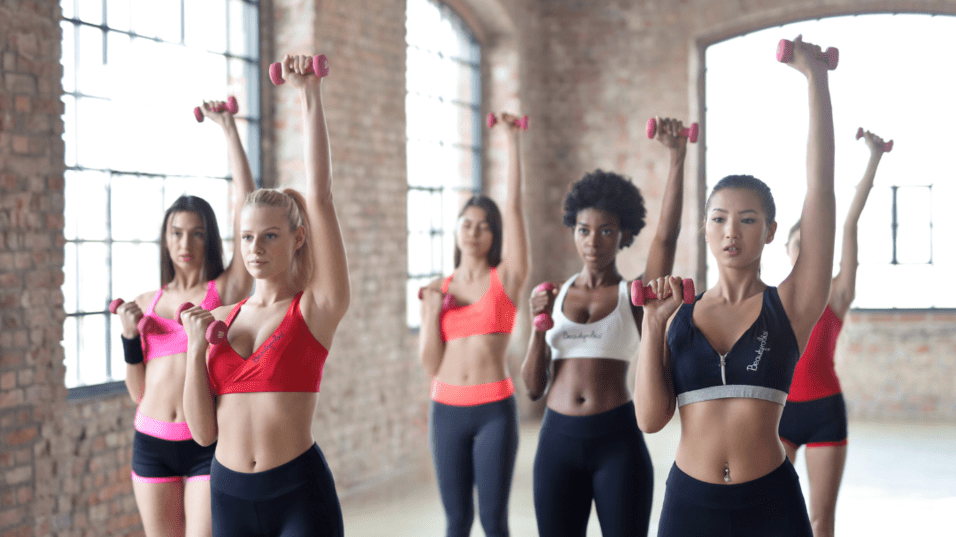 Gruppe von sechs Frauen in Sportbekleidung, die Hanteln in einer hellen, industriellen Umgebung heben.