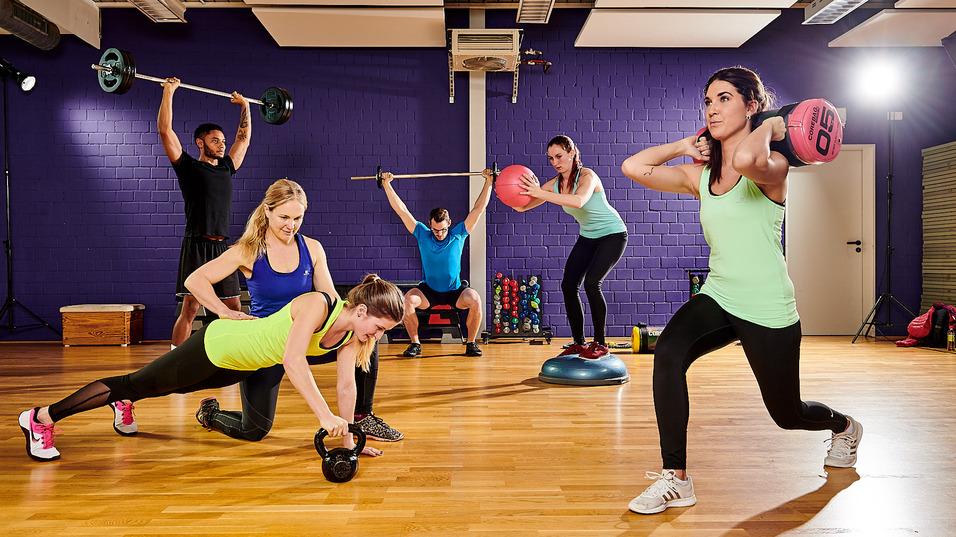 Gruppenfitness: Sechs Personen trainieren gemeinsam im Gym mit Gewichten und Fitnessgeräten auf einem Holzboden.