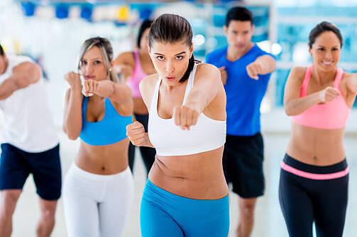 Gruppe von sechs Menschen in Sportkleidung, die synchron F&auml;uste aussto&szlig;en w&auml;hrend eines Fitnesskurses in einem Studio.