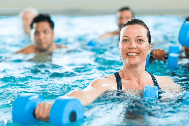 Frau mit blauen Schwimmhanteln im Wasser, l&auml;chelt gl&uuml;cklich w&auml;hrend eines Aquafitness-Kurses, andere Teilnehmer im Hintergrund.