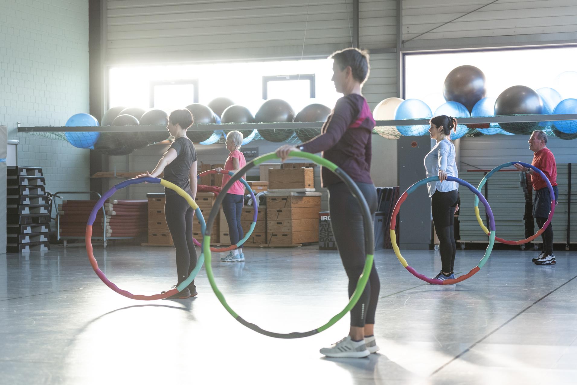 Gruppe von fünf Personen im Fitnessstudio mit Hula-Hoops in einer Bewegungsübung bei Sonnenlicht.