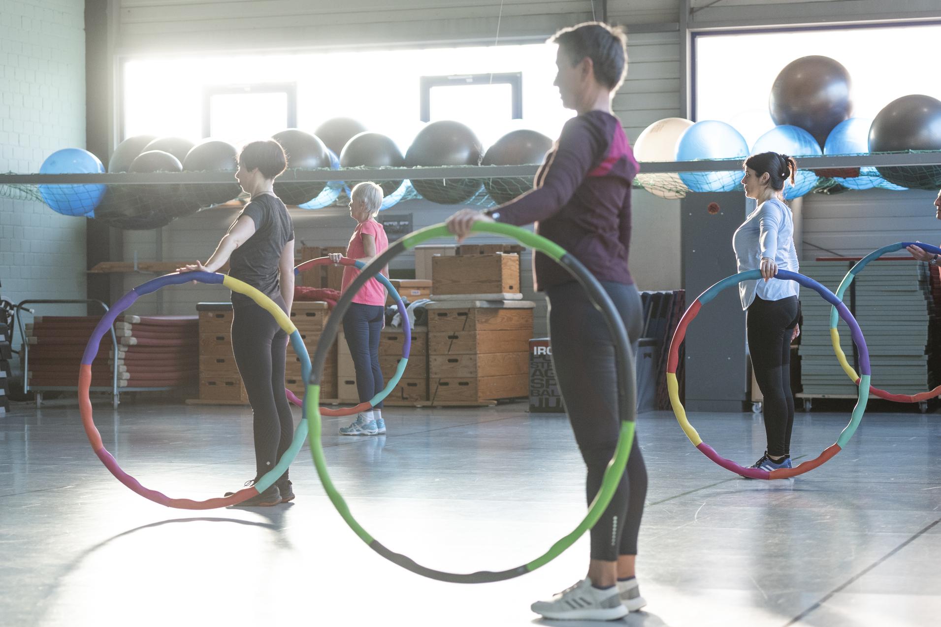 Gruppe von f&uuml;nf Personen im Fitnessstudio mit Hula-Hoops in einer Bewegungs&uuml;bung bei Sonnenlicht.