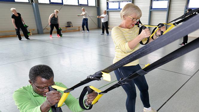 Gruppe von Menschen unterschiedlichen Alters trainiert mit TRX-Bändern in einem lichtdurchfluteten Gymnastikraum.