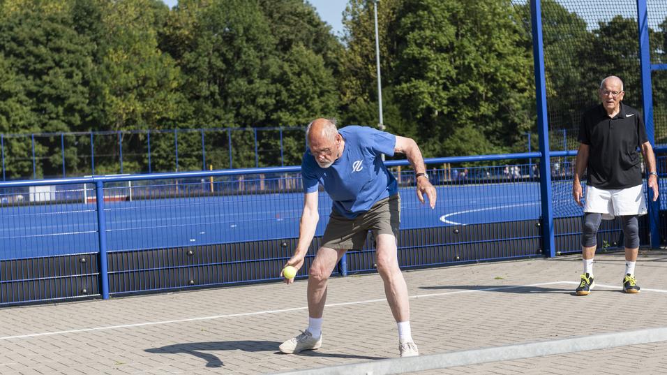 Zwei Männer auf einem Sportplatz, einer schlagt einen Ball, der andere steht wartend im Hintergrund.