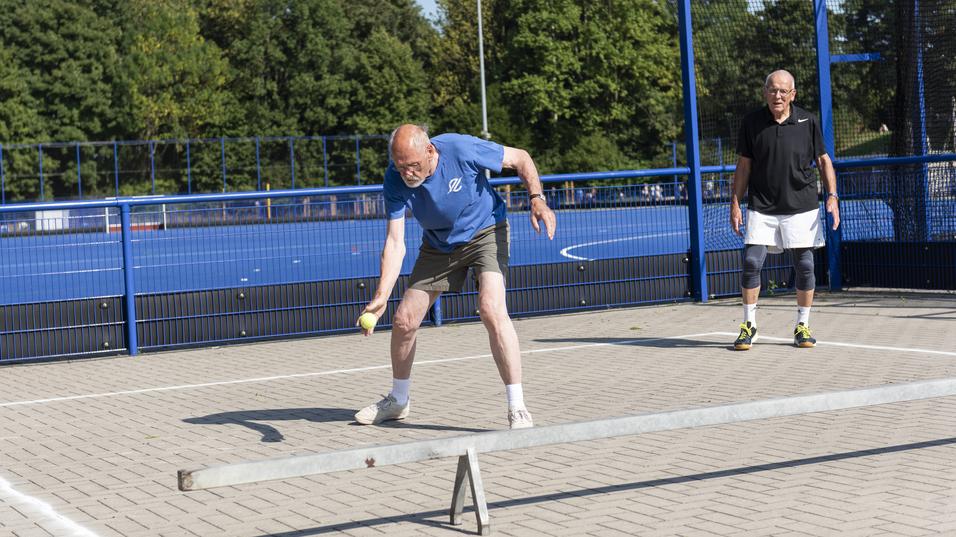 Zwei M&auml;nner auf einem Sportplatz, einer schlagt einen Ball, der andere steht wartend im Hintergrund.
