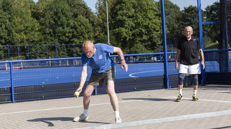 Zwei M&auml;nner auf einem Sportplatz, einer schlagt einen Ball, der andere steht wartend im Hintergrund.